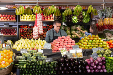 Young male vendor in an apron, in the department with fresh vegetables and fruits in the supermarket, a woman in the background