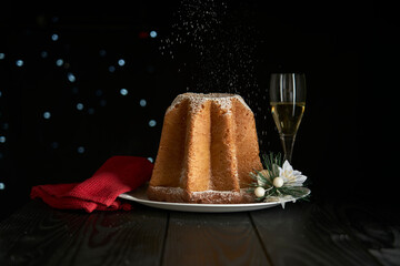 Celebrating Christmas with a snowfall of powdered sugar that whitens a pandoro, a typical dessert of the Christmas tradition, with a glass of sparkling wine and a bokeh effect on a dark background.