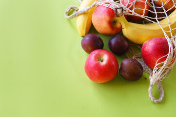 Red apples, bananas, plums and string bag on a bright green background.