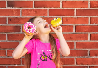 A girl with bright donuts in her hands is making faces against the background of a brick wall.
