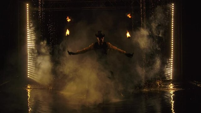A male fire tamer spins flaming balls of fire on chains while standing among the raindrops. An exciting fire show in a dark studio at night. A silhouette in the smoke. Slow motion.
