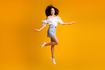 Fototapeta na wymiar Photo portrait of jumping high girl curly hairstyle wearing jeans mini skirt sneakers laughing isolated on vivid yellow color background