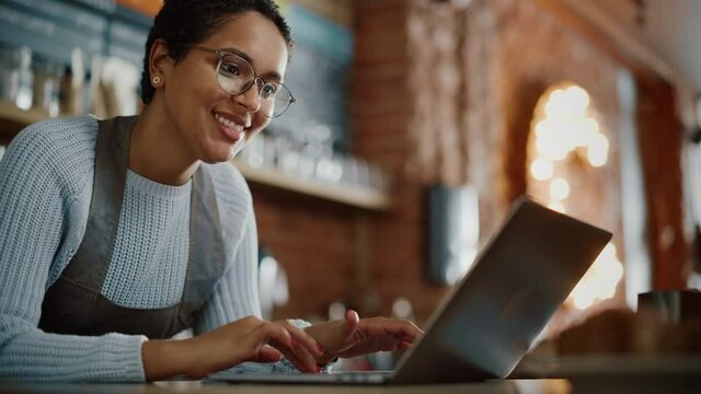 Young and Beautiful Latina Coffee Shop Owner is Working on Laptop Computer and Checking Inventory in a Cozy Cafe. Happy Restaurant Manager or Employee Browsing Internet and Chatting with Friends.