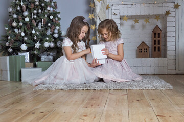 Christmas and new year decorated card. Two girls opening giftbox near fireplace. Sisters in beautiful party dresses sitting on the floor new year background