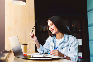 Attractive business woman is sitting at table in front of laptop. Beautiful brunette girl smiles, holds glasses