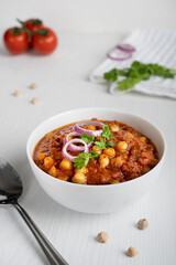 Chole masala or chana an indian street food made of chickpeas, tomatoes and cumin decorated with onion rings and parsley served in bowl with spoon on white wooden background. Vertical image