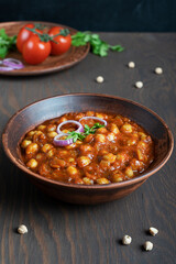Chole masala or chana an indian street food made of chickpeas, tomatoes and cumin decorated with onion rings and parsley served in bowl at kitchen on dark brown wooden background. Vertical image
