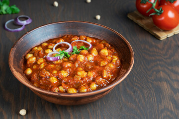 Chole masala or chana an indian street food made of chickpeas, tomatoes and cumin decorated with red onion rings and parsley served in bowl at kitchen on dark brown wooden background. Horizontal image