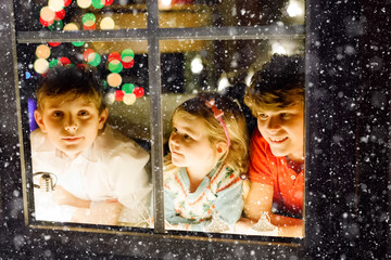 Three cute children sitting by window on Christmas eve. Two school kid boys and toddler girl, siblings looking outdoor and dreaming. Family happiness on traditional holiday
