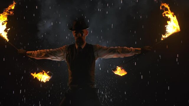 A male fire tamer in a spectacular close up fire show against a dark studio background among raindrops. A silhouette of a man in smoke waves flaming torches. Slow motion. Close up.