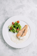 A piece of baked salmon with sour cream sauce with green salad on a white plate top view