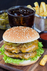 Double cheese burger with two beef patty, cheddar cheese, lettuce, caramelized onion and jalapeno slices, served with french fries and soda, on wooden board, vertical, closeup