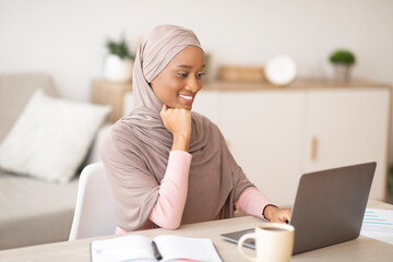 Happy black Muslim woman in hijab working remotely on laptop from home