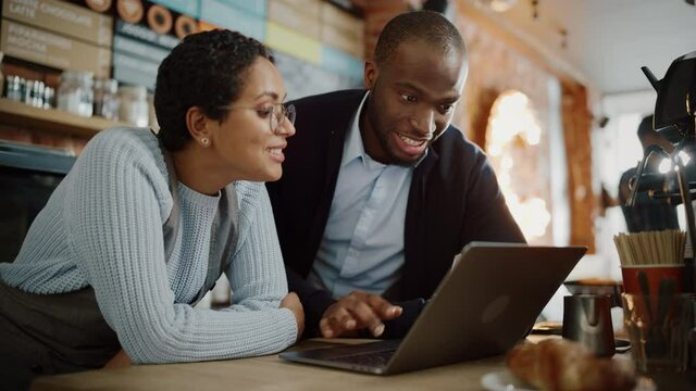 Two Diverse Entrepreneurs Have a Team Meeting in Their Stylish Coffee Shop. Barista and Cafe Owner Discuss Work Schedule and Menu on Laptop Computer. Multiethnic Female and Male Restaurant Employees.