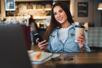 Close-up. Beautiful smiling young business woman sits in front of laptop, holds cup of coffee, uses smartphone for work
