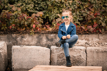 A cute little girl is sitting in sunglasses on a stone fence. Stylish child in a blue jacket. Casual style, fashion for children, fashionable suit, happy childhood