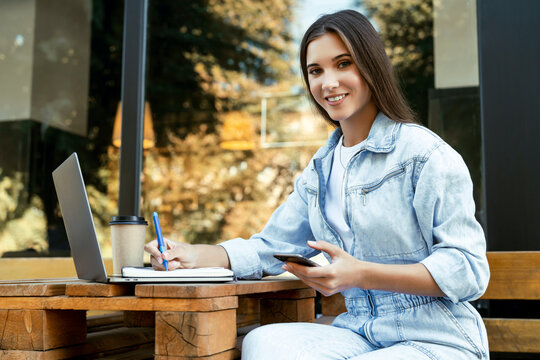 Young business woman working on open terrace at home, sitting in front of laptop, holding smartphone in her hand. 