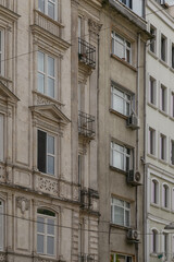 Fototapeta na wymiar Old building facade with windows decorated with stucco frontones, false columns and balconies with wrought-iron bars. Vertical photo of a building with elements of city ​​communications.