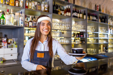 Beautiful female barista is holding a cup with hot coffee, wearing face shield while standing near the bar counter in cafe. New normal