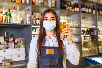 Beautiful female Bartender wearing protective face mask, serving a draft beer during coronavirus pandemic, shelves full of bottles with alcohol on the background