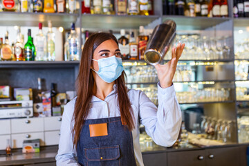 Young female worker at bartender desk in restaurant bar preparing coctail with shaker. Beautiful young woman behind bar wearing protective face mask