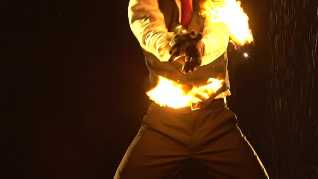 Male fire tamer in a thrilling close up fire show in a dark studio in the rain. A silhouette of a man swinging fiery balls on chains in slow motion.