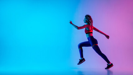 Fototapeta na wymiar Runner. Young sportive woman training isolated on gradient blue-pink studio background in neon light. athletic and graceful. Modern sport, action, motion, youth concept. Beautiful female practicing.