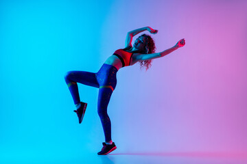 Naklejka na ściany i meble Sport dancing. Beautiful woman in bright sportwear isolated on gradient pink-blue background in neon light. Athletic and graceful. Modern sport, action, motion, youth concept. Female practicing
