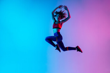 Jump. Young beautiful woman in bright sportwear isolated on gradient pink-blue background in neon...