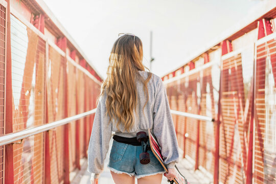 Beautiful blonde young woman walking away on a bridge holding a skateboard. Cool female model from the back. Urban California style outfit. 