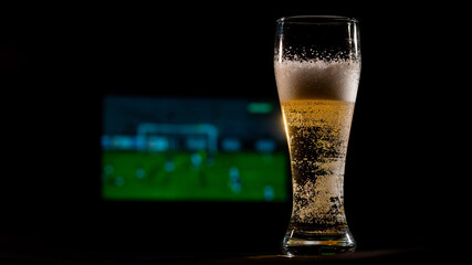 Close-up of fresh foaming light beer in a glass and football on TV on background. Sports bar concept
