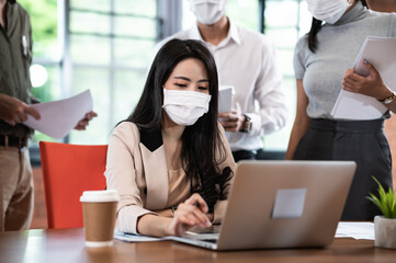 Attractive Asian female boss supervisor wear protective face mask in New normal office teaching and explaining intern or new employee to use corporate software or helping with difficult assignment