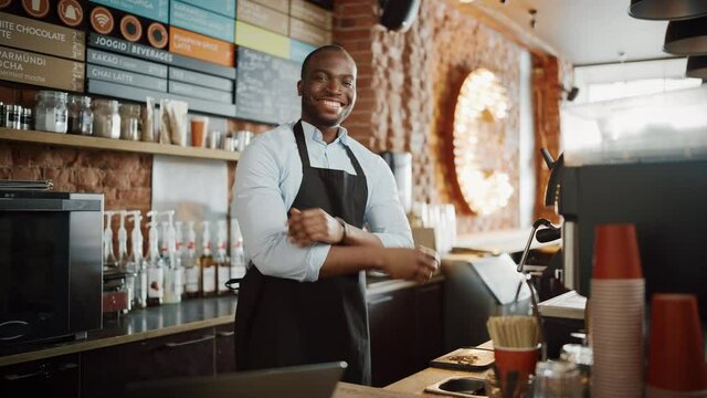 Handsome Black Afro-American Barista Wearing Apron is Making a Cup of Tasty Cappuccino in Coffee Shop Restaurant. Portrait of Happy Employee Behind Cozy Loft-Style Cafe Counter.