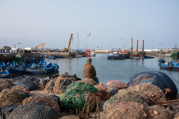 homme assis sur des filets de pêche face à un port de pêche 