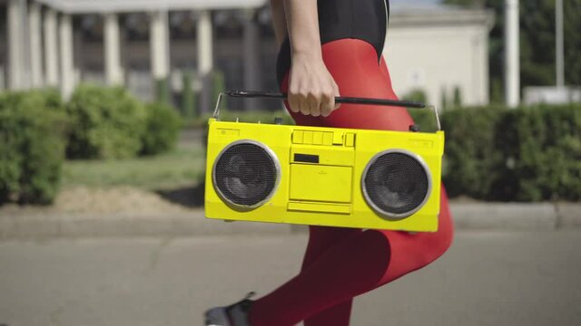 Unrecognizable slim woman in red legging walking on sunny summer street with vintage yellow tape recorder. Fit Caucasian 1980s sportswoman strolling and dancing to music outdoors.