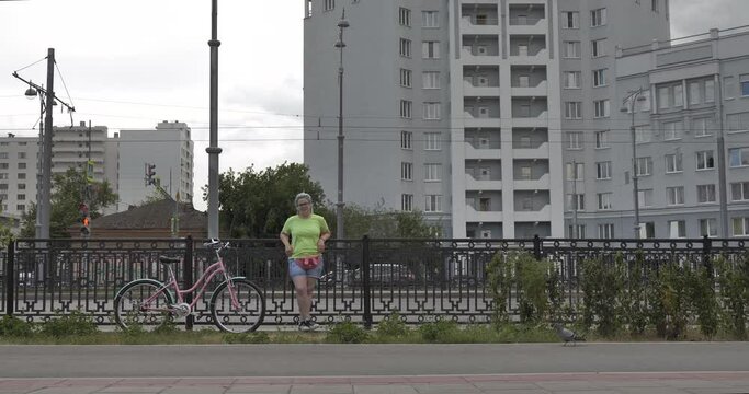 The general shot of a bright plus size girl, she is leaning on the city fence and looking out into the street. Her pink bike is parked next to the heroine.