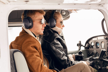 Beautiful, young, stylish couple in winter clothes, glasses and headphones, sitting in the cockpit of a small pleasure plane.