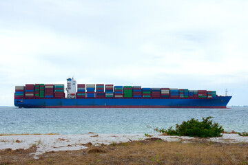 Large colorful container shipping tanker boat vessel loaded with steel storage boxes cargo cruising close by in the water by a beach on a calm morning.