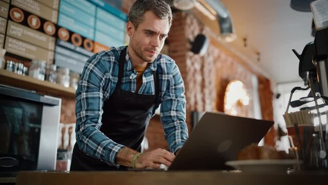 Young and Handsome Coffee Shop Owner is Working on Laptop Computer and Checking Inventory in a Cozy Cafe. Happy Restaurant Manager in Checkered Shirt Browsing Internet and Chatting with Friends.