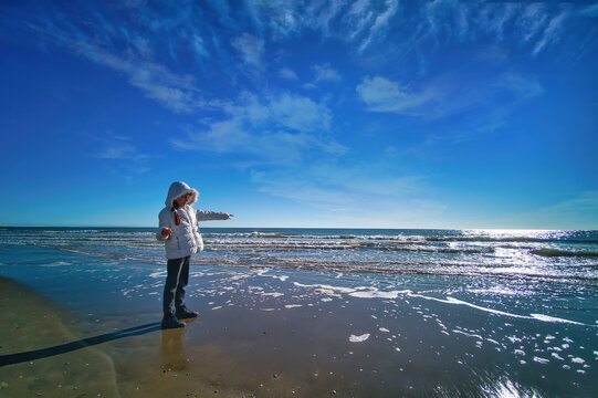 Bambina al mare in inverno in una bella giornata di sole, mentre indica all'orizzonte vestita di bianco