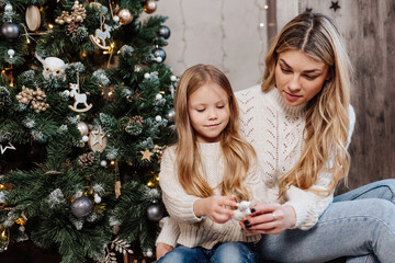 Young mother and her cute daughter sitting near Christmas tree indoors.