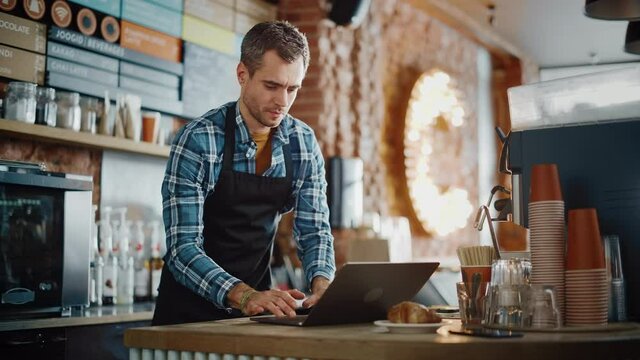Young and Handsome Coffee Shop Owner is Working on Laptop Computer and Checking Inventory in a Cozy Cafe. Happy Restaurant Manager in Checkered Shirt Browsing Internet and Chatting with Friends.