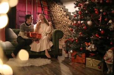 Fototapeta na wymiar long-haired young girl and a guy sit on the sofa next to a christmas tree, boxes with gifts, festive home decor, new year atmosphere, selective focus, bokeh