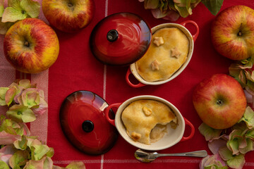 Naklejka na ściany i meble Little apple pies in pots with bright red lids on a red table cloth, decorated with apples and flowers.