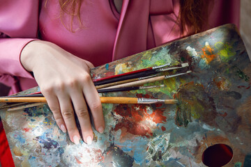 woman in a pink jacket stands with paints, brushes and palette on the street
