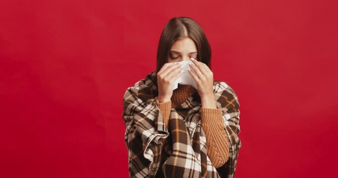 Young lady wrapped in plaid sneezing, blowing her nose into tissue, got sick before holidays, red studio background
