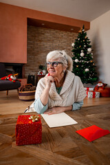 Obraz na płótnie Canvas Senior smiling woman, sitting on the floor at home, thinking what to write in a blank paper. Grey hair lady with greeting cards, wrapped gifts and christmas tree. Alone for Christmas.