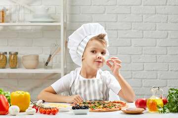 child making tasty delicious gesture by kissing fingers. little boy in chef hat and an apron...