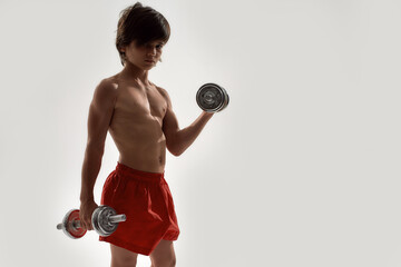 Little sportive boy child with muscular body looking at camera, showing his muscles, lifting weights while standing isolated over white background