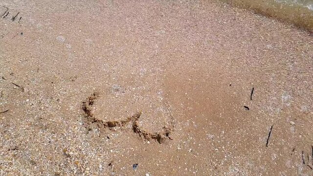 The wave washes away the heart nestled in the sand. Woman draws a heart in the sand. A woman draws a heart on the seashore which is washed away by a wave.